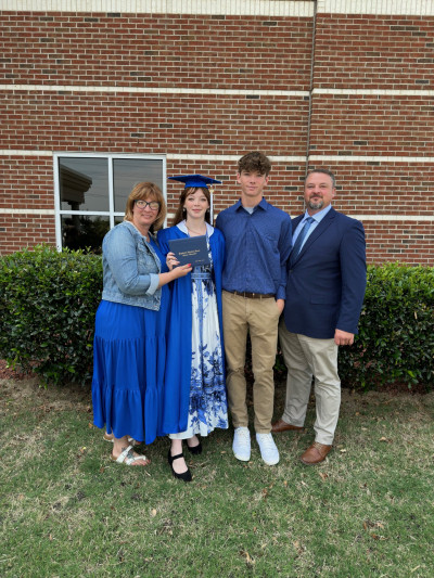 Lilly Guinn stands in blue cap and graduation gown, holding her diploma smiling alongside her mother, father, and brother.